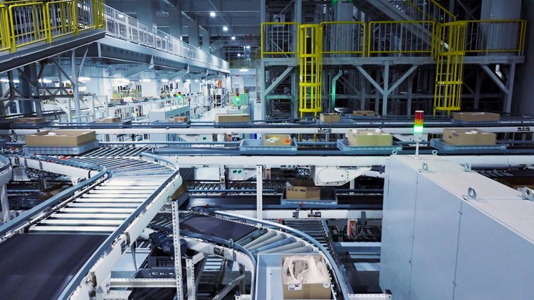 In einer großen Lagerhalle sind Förderbänder zu sehen, die Kartons transportieren. Mehrere Ebenen und Ausstattungsteile deuten auf ein automatisiertes Logistiksystem hin. Beleuchtung und Sicherheitsvorrichtungen sind vorhanden.