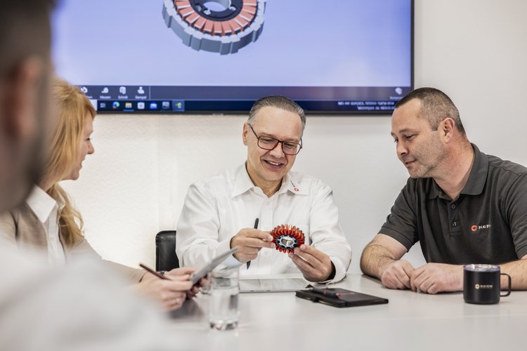 Teammeeting mit Produktpräsentation Vier Personen in einem Besprechungsraum, ein Mann in weißem Hemd präsentiert ein technisches Bauteil, im Hintergrund ein Bildschirm mit Firmenlogo.